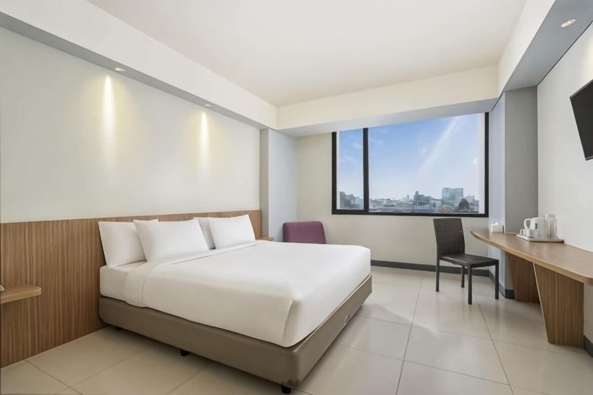 A modern hotel room with a large bed, a desk, a chair, and a city view.