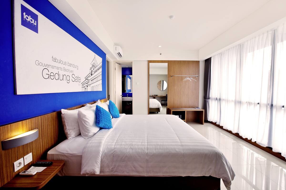 A modern hotel room with a large bed, a nightstand, a desk, and a bathroom visible in the background.