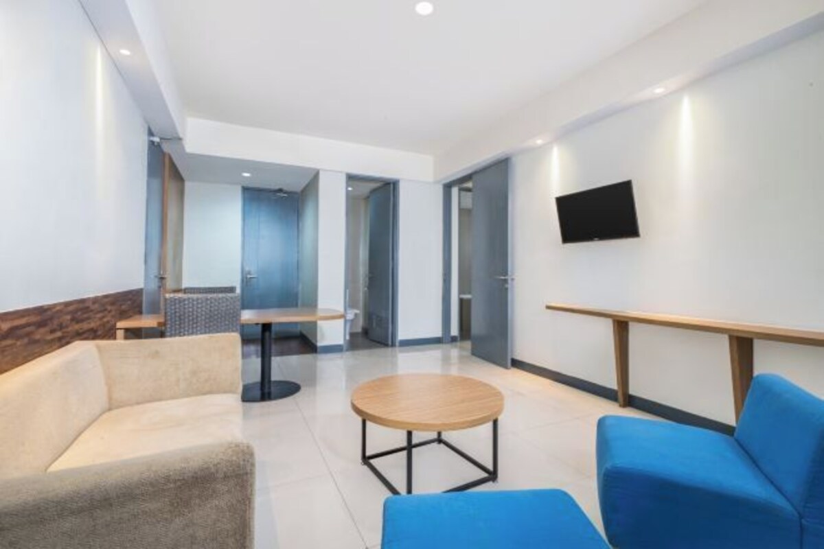 Um hall de hotel moderno com uma mesa redonda, cadeiras azuis, um sofá e uma televisão de tela plana fixada na parede.