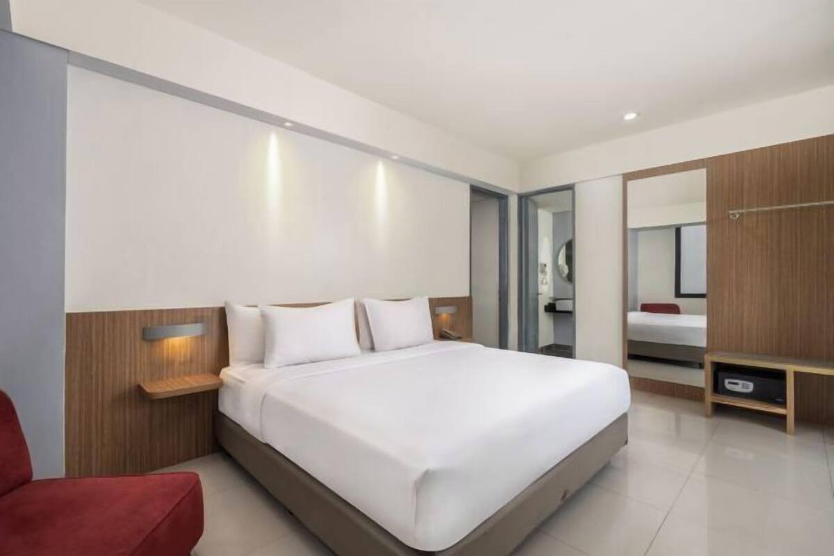 Quarto de hotel moderno com uma cama grande, uma cadeira vermelha, uma mesa de cabeceira de madeira e um espelho.