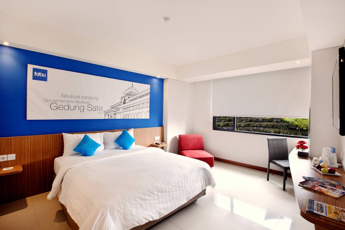 A modern hotel room with a large bed, a red chair, a desk with a computer, and a window with a view of greenery.