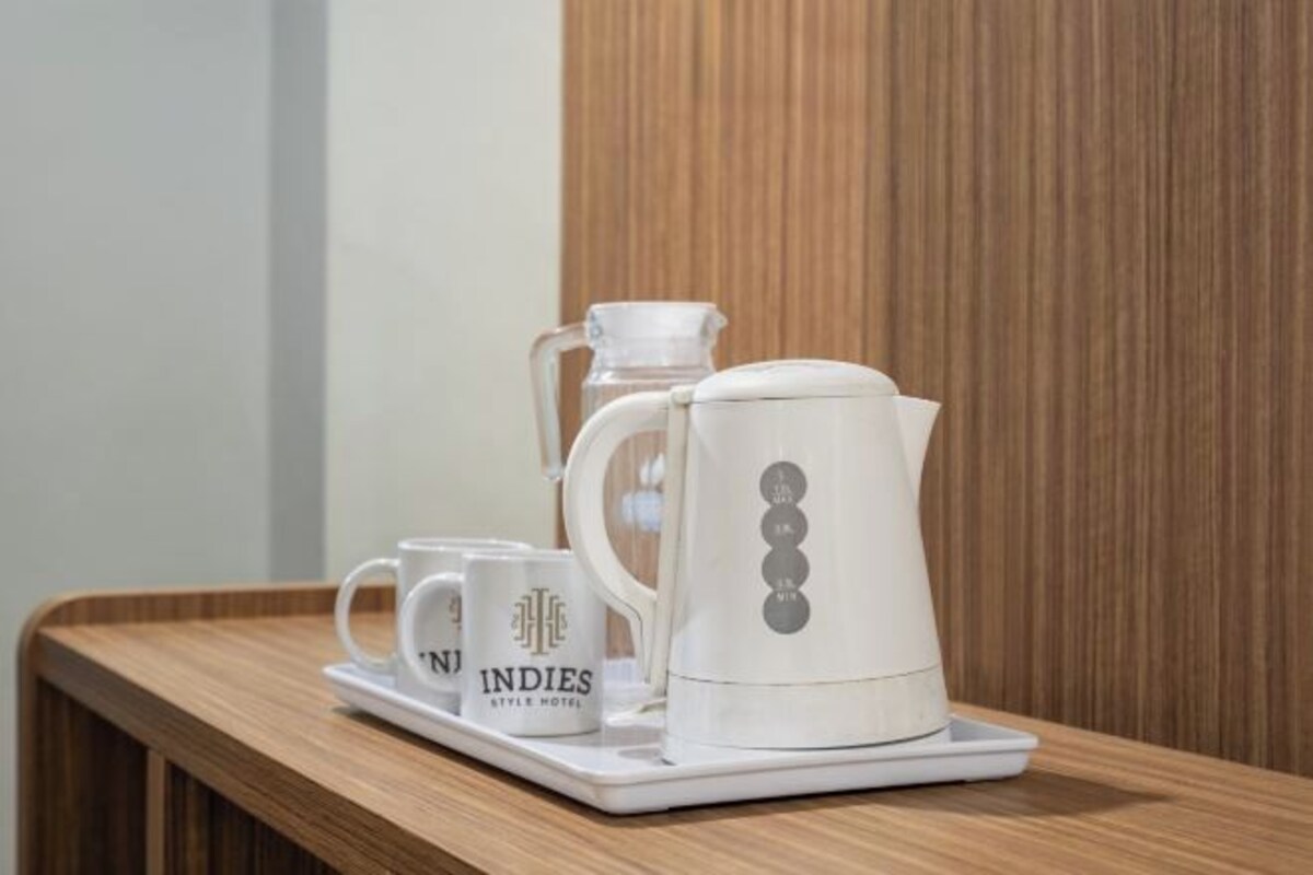 Máquina de café branca, jarra de vidro e duas chávenas brancas com a inscrição "INDIES STYLE HOTEL" sobre um tabuleiro de madeira.