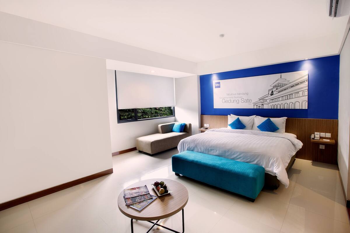 A modern hotel room with a large bed, a small sofa, a round table with magazines, and a wall-mounted artwork.