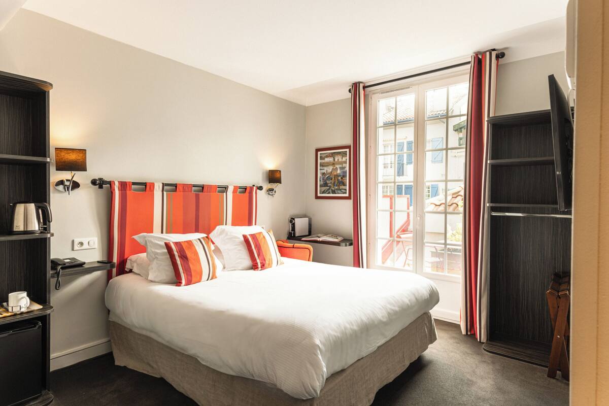 Ein Hotelzimmer mit einem großen Bett, einem Schreibtisch mit Lampe, einem Bücherregal und einem Fenster mit Vorhängen.