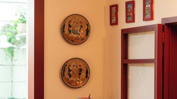 Canto de um quarto com uma estátua de sapo decorativo sobre um banquinho, uma janela com persianas vermelhas e duas imagens emolduradas na parede.