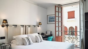 A hotel room with a bed, a desk, a chair, a window with curtains, and a balcony with a railing. - Boutique Hôtel Txoko (Saint-Jean-de-Luz)