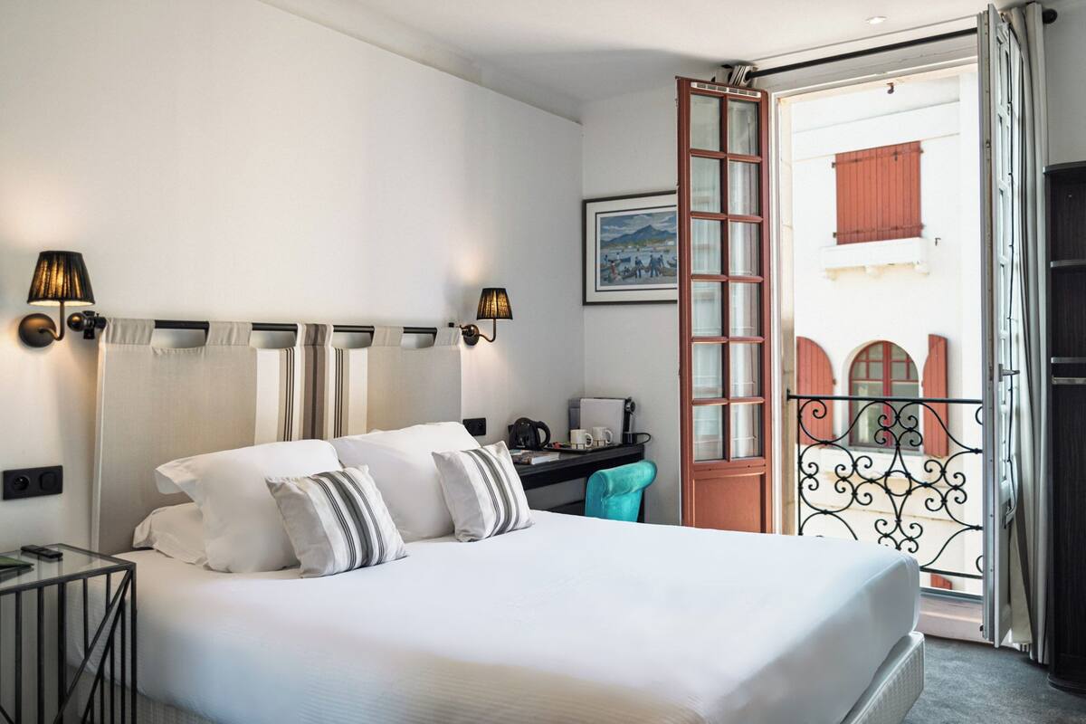 Ein Hotelzimmer mit Bett, Schreibtisch, Stuhl, einem Fenster mit Vorhängen und einem Balkon mit Geländer.