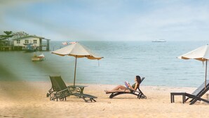 Une plage avec des chaises longues et des parasols, une femme qui lit un livre et un bateau sur l’eau.