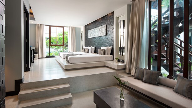 Une chambre moderne avec un grand lit, un canapé et une table basse. Un escalier mène à un balcon offrant une vue sur la verdure.