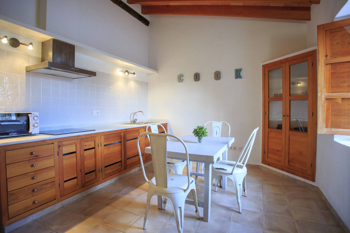 Una cocina con armarios de madera, encimera blanca, microondas y una mesa de comedor con sillas.