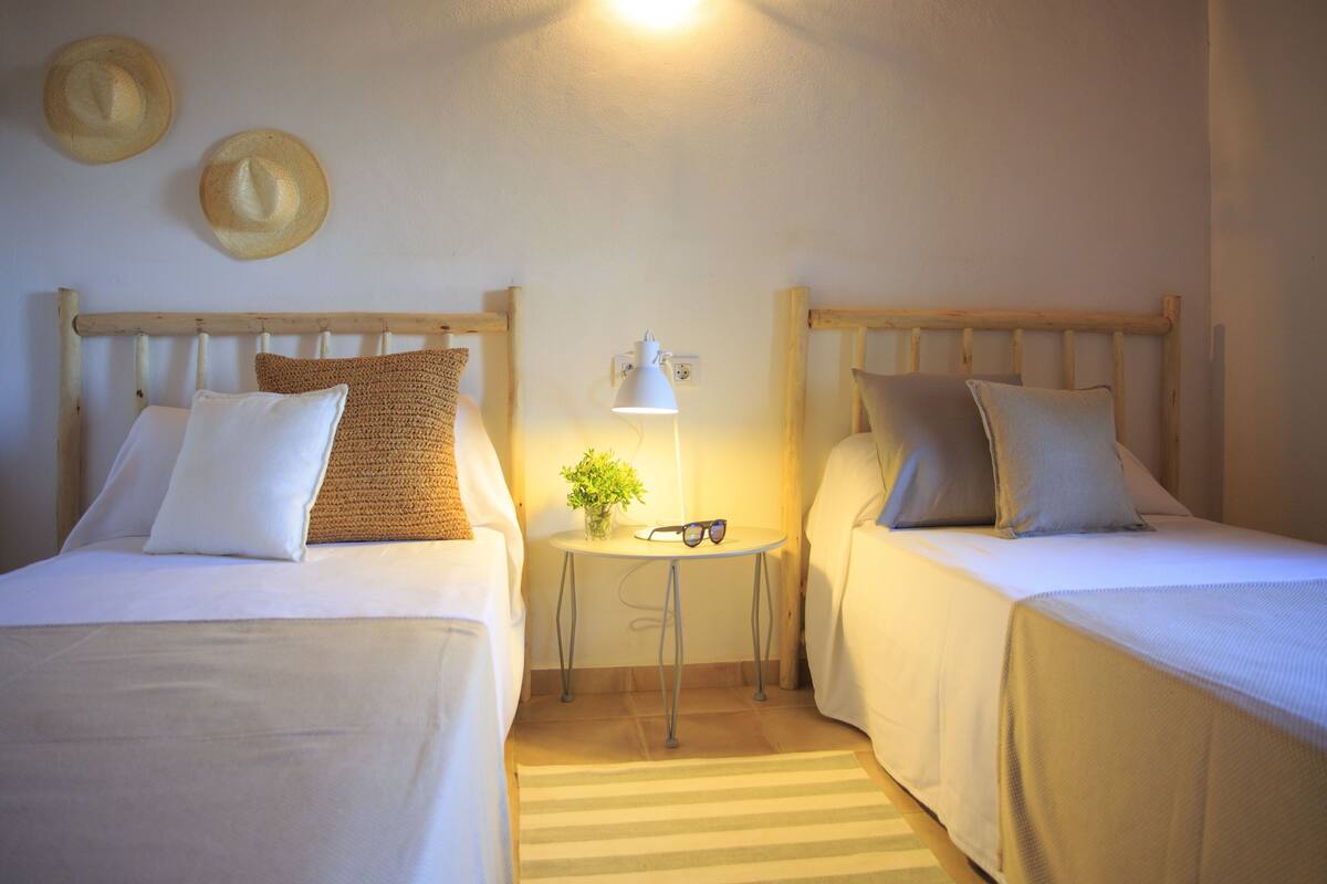 Habitación con dos camas, cada una con cabecero de madera, ropa de cama blanca y una pequeña mesa con una lámpara y una planta entre ellas.