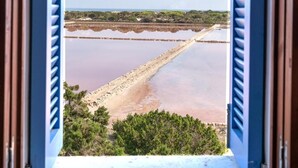 Une vue à travers une fenêtre ornée de volets bleus, donnant sur une plage où la mer est calme et où l’on aperçoit une côte lointaine.