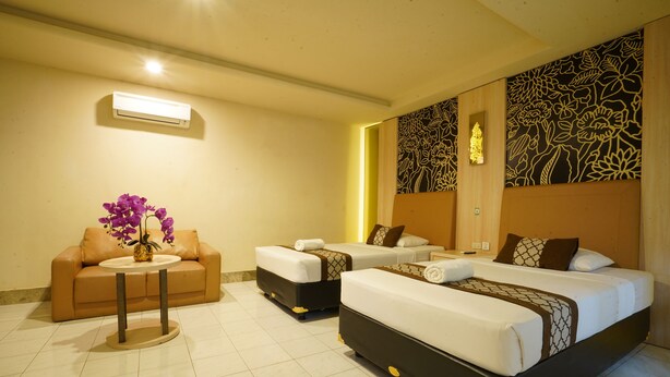 A hotel room with two beds, a sofa, a small table with a vase of flowers, and a decorative wall panel. - Hotel GRAND ROSELA Yogyakarta (Yogyakarta)
