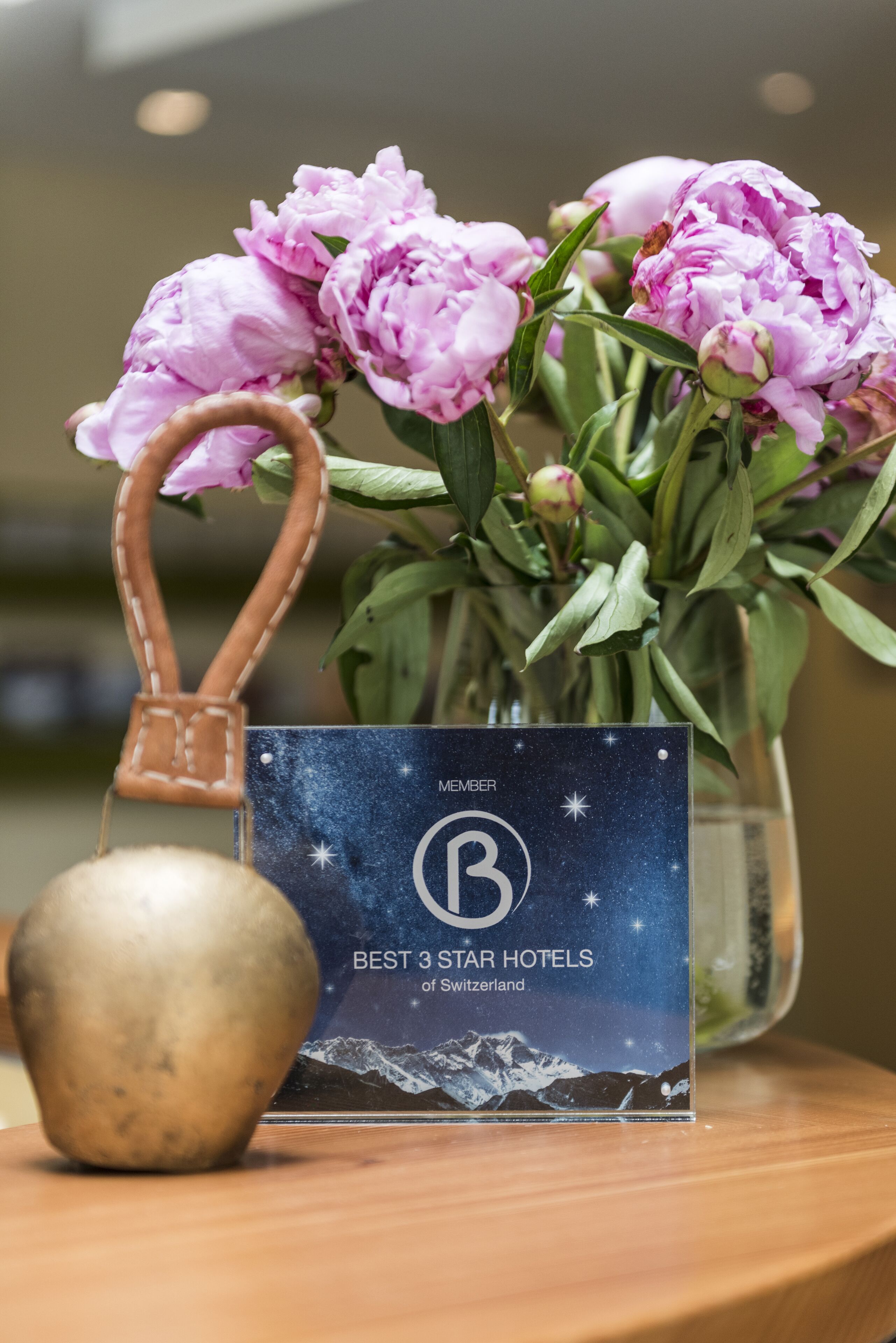 Un bouquet de pivoines roses dans un vase, une cloche en laiton et une carte répertoriant les trois meilleurs hôtels trois étoiles de Suisse.