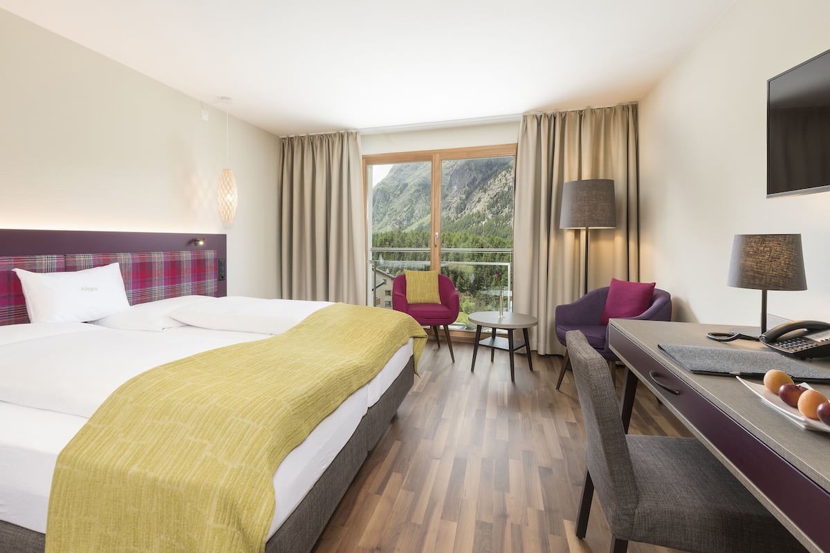 Een hotelkamer met een bed, een bureau, een stoel, een klein tafeltje en uitzicht op bergen door het raam.