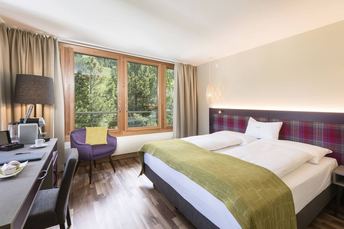 Hotelkamer met een bed, een bureau met computer, een stoel en een raam met uitzicht op bomen.