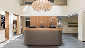 Un espace d’accueil d’hôtel avec un comptoir de réception en bois, un grand luminaire suspendu et une enseigne indiquant le nom de l’hôtel.
