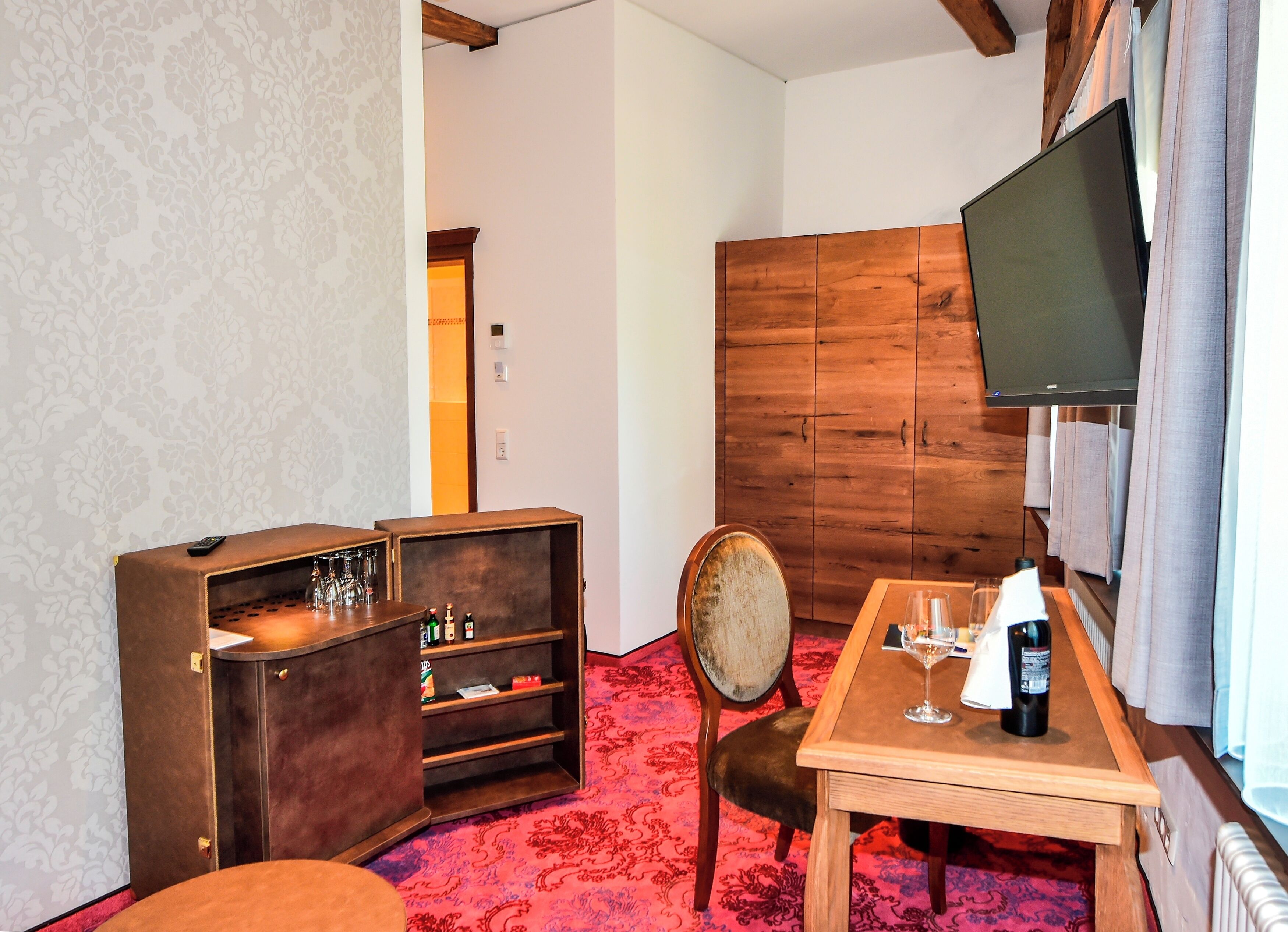 Habitación de hotel con un armario de madera, un televisor, una mesa de comedor con una silla y una alfombra estampada.