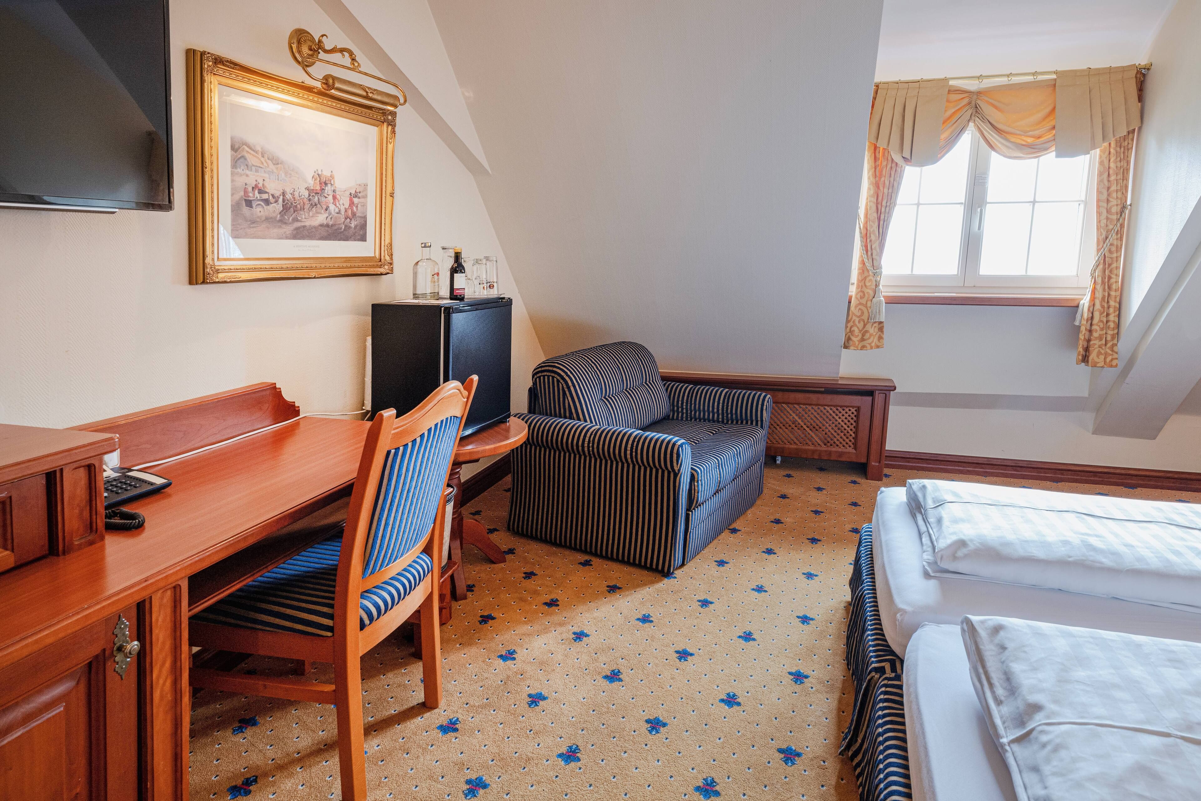 Habitación de hotel con un escritorio de madera, un sillón a rayas, una cama con sábanas blancas, un televisor y un cuadro en la pared.