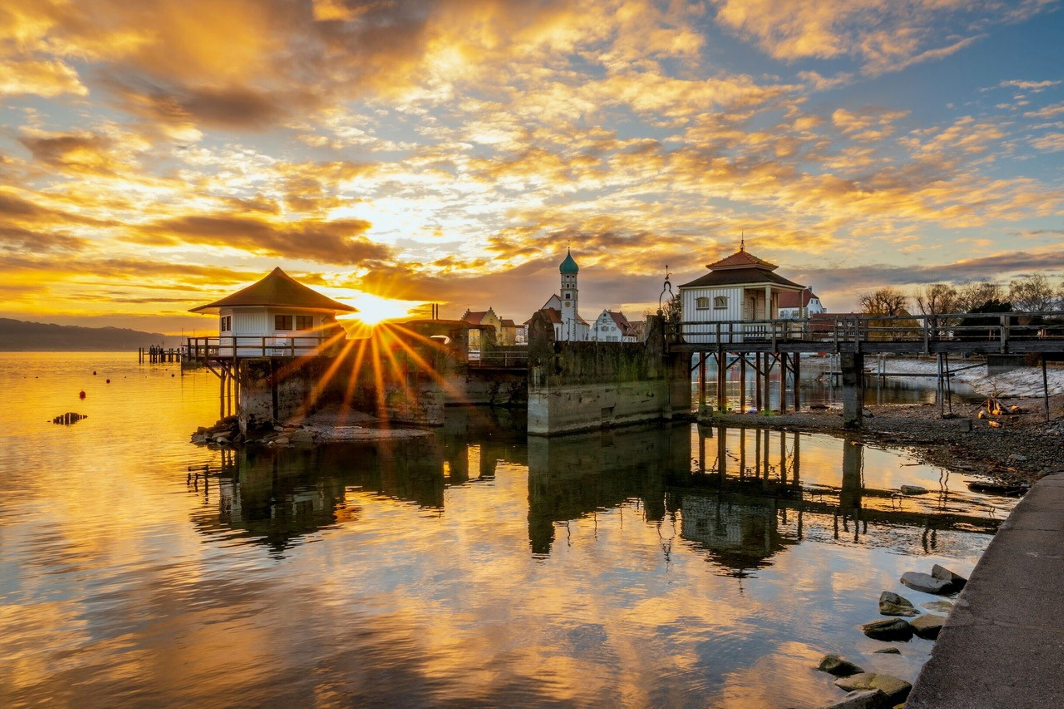 Escena costera con edificios y un muelle reflejando la luz del atardecer.