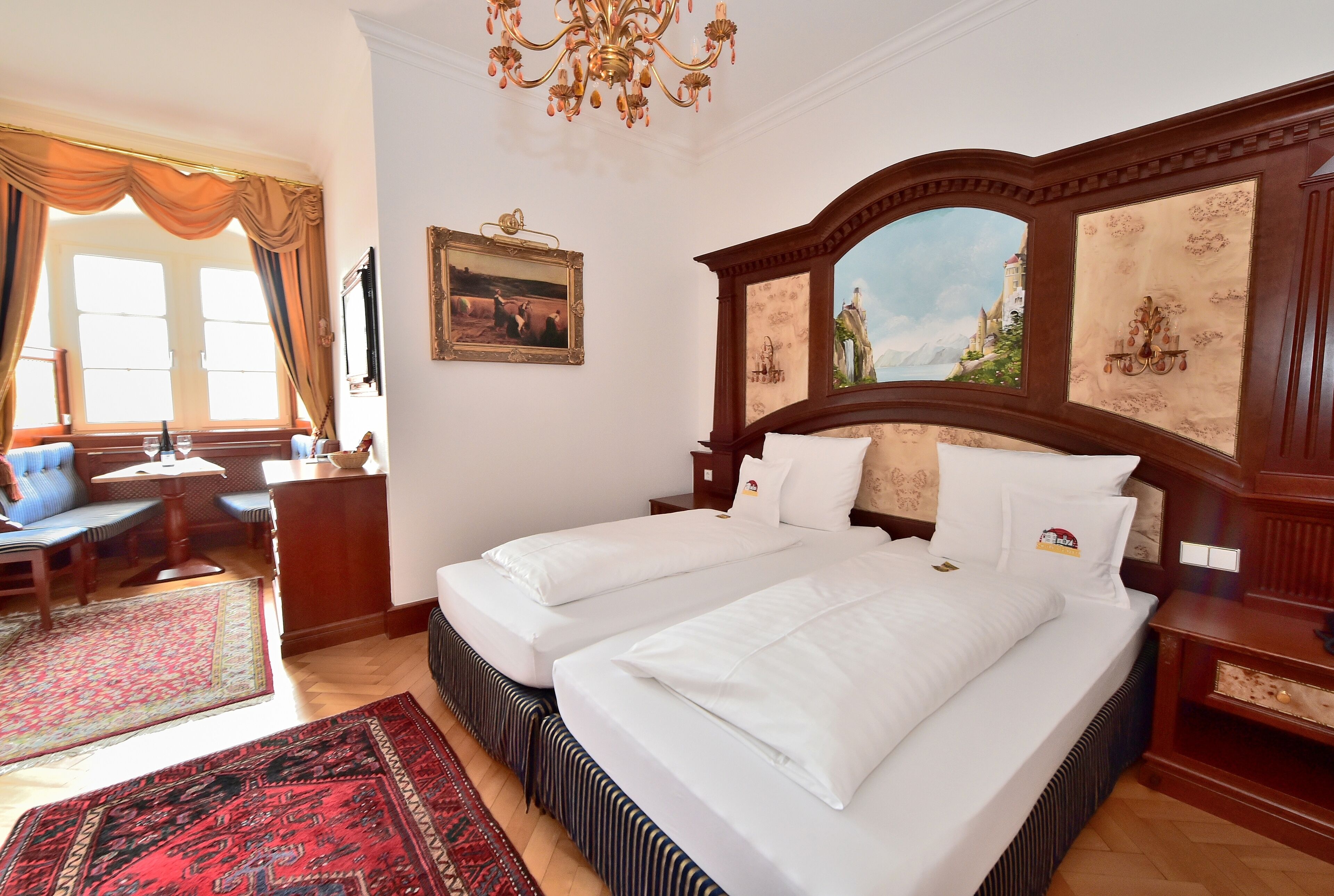 Une chambre d’hôtel avec deux lits, un coin salon avec un canapé et des fauteuils, une grande fenêtre avec des rideaux et un lustre.