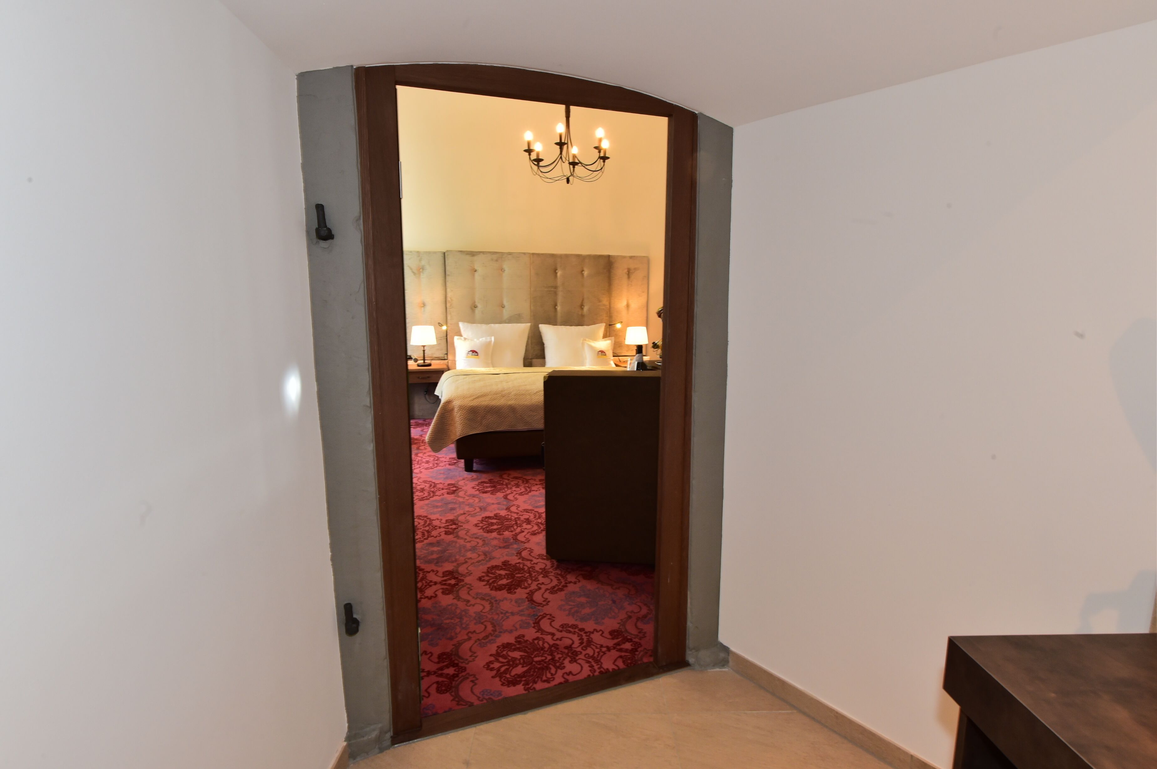 Una habitación de hotel con un arco de entrada que conduce a un dormitorio con cabecera acolchada, dos mesitas de noche con lámparas y una alfombra roja con estampado.