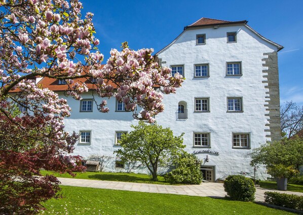 A white building with multiple windows and a red roof, surrounded by green grass and flowering trees. - Schloss Hotel Wasserburg (Wasserburg am Bodensee)