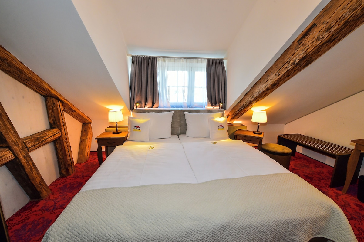 A bedroom with a large bed, two bedside tables with lamps, a window with curtains, and wooden beams on the ceiling.
