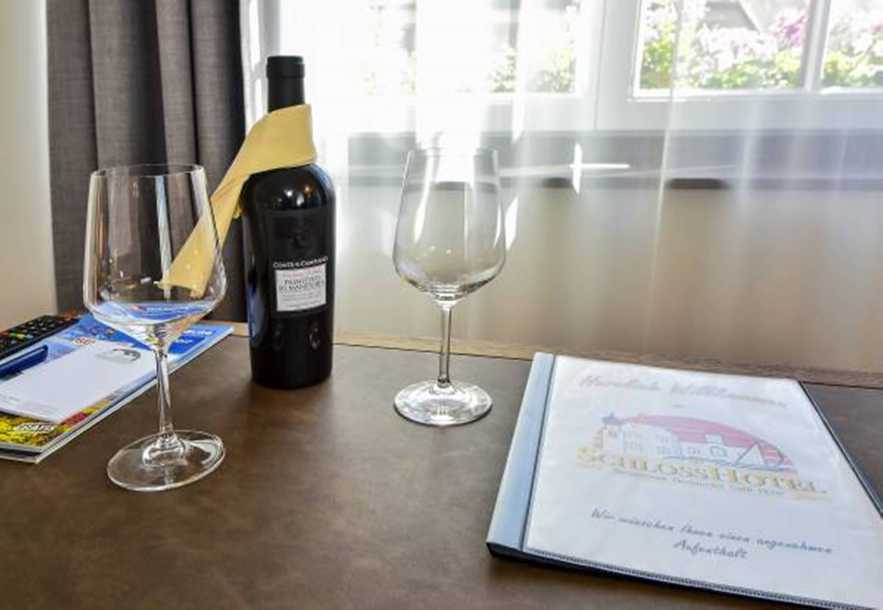 A table with a bottle of wine, two wine glasses, a menu, and a booklet on a wooden table near a window.