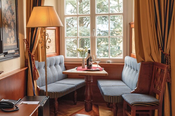 A corner seating area with two striped cushioned benches, a small table with a vase and wine glasses, and a lamp. - Schloss Hotel Wasserburg (Wasserburg am Bodensee)