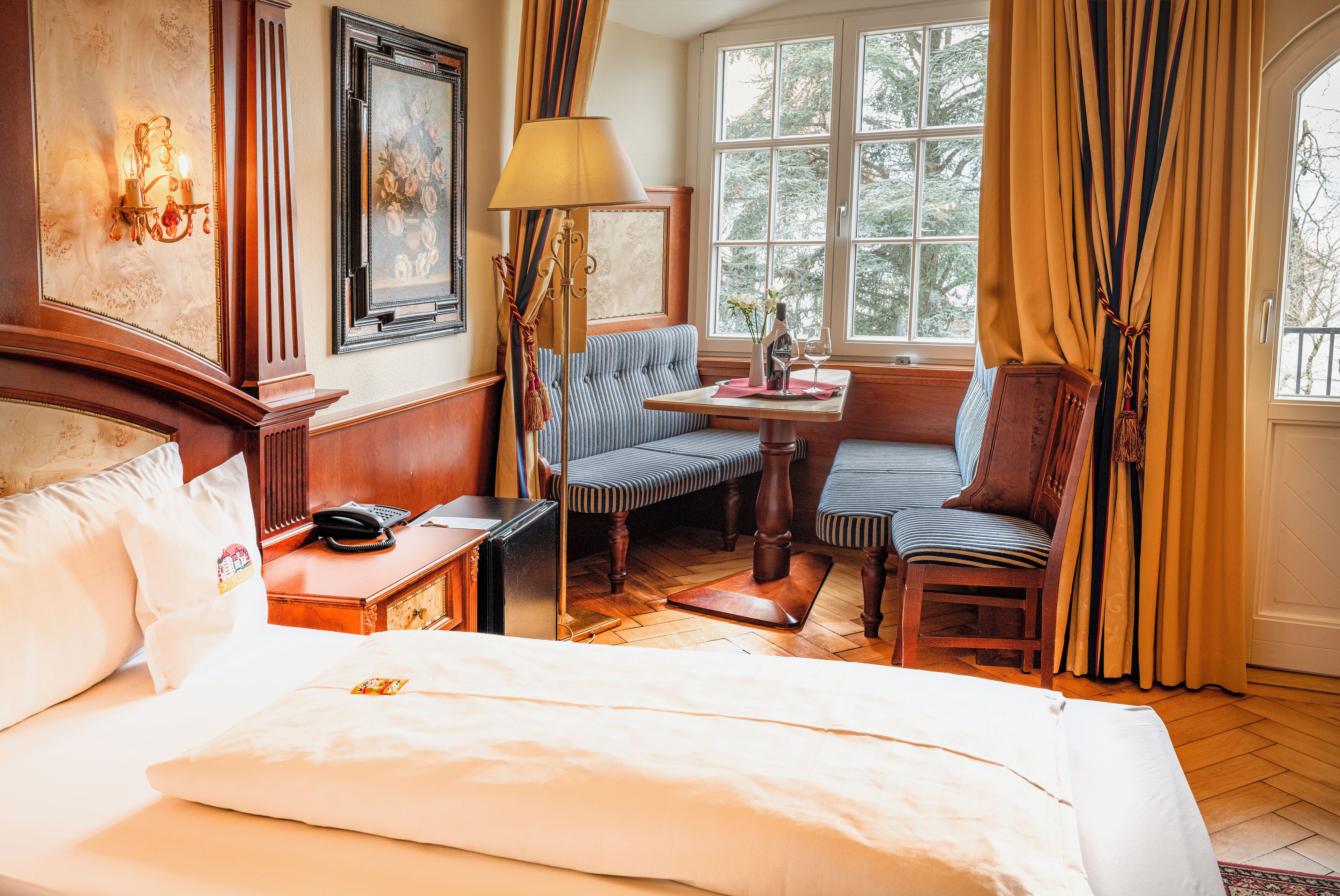 A hotel room with a bed, a desk with a telephone, a chair, a table with a vase and wine glass, and a window with curtains.