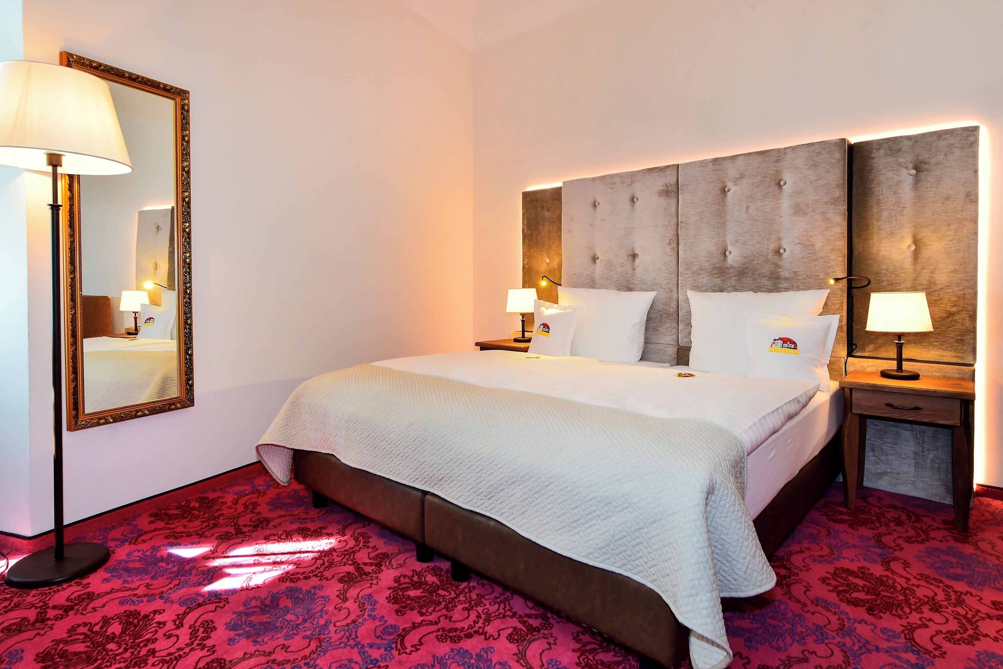 Una habitación de hotel con una cama grande, una mesita de noche, un espejo y una alfombra roja estampada.