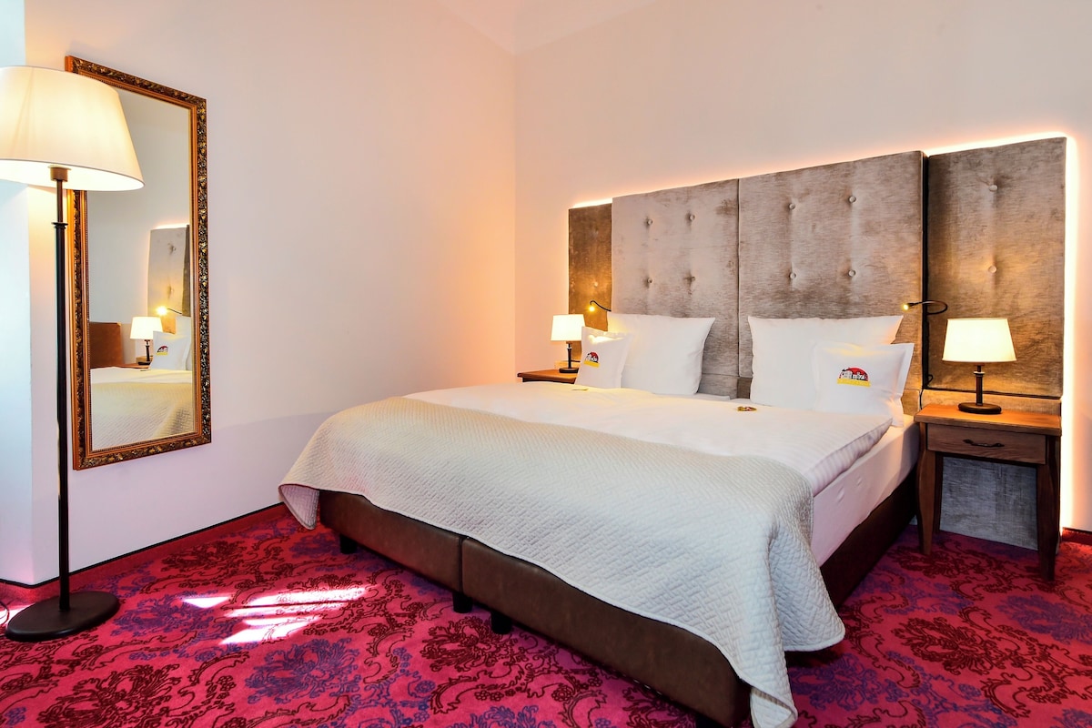 A hotel room with a large bed, a nightstand, a mirror, and a red patterned carpet.