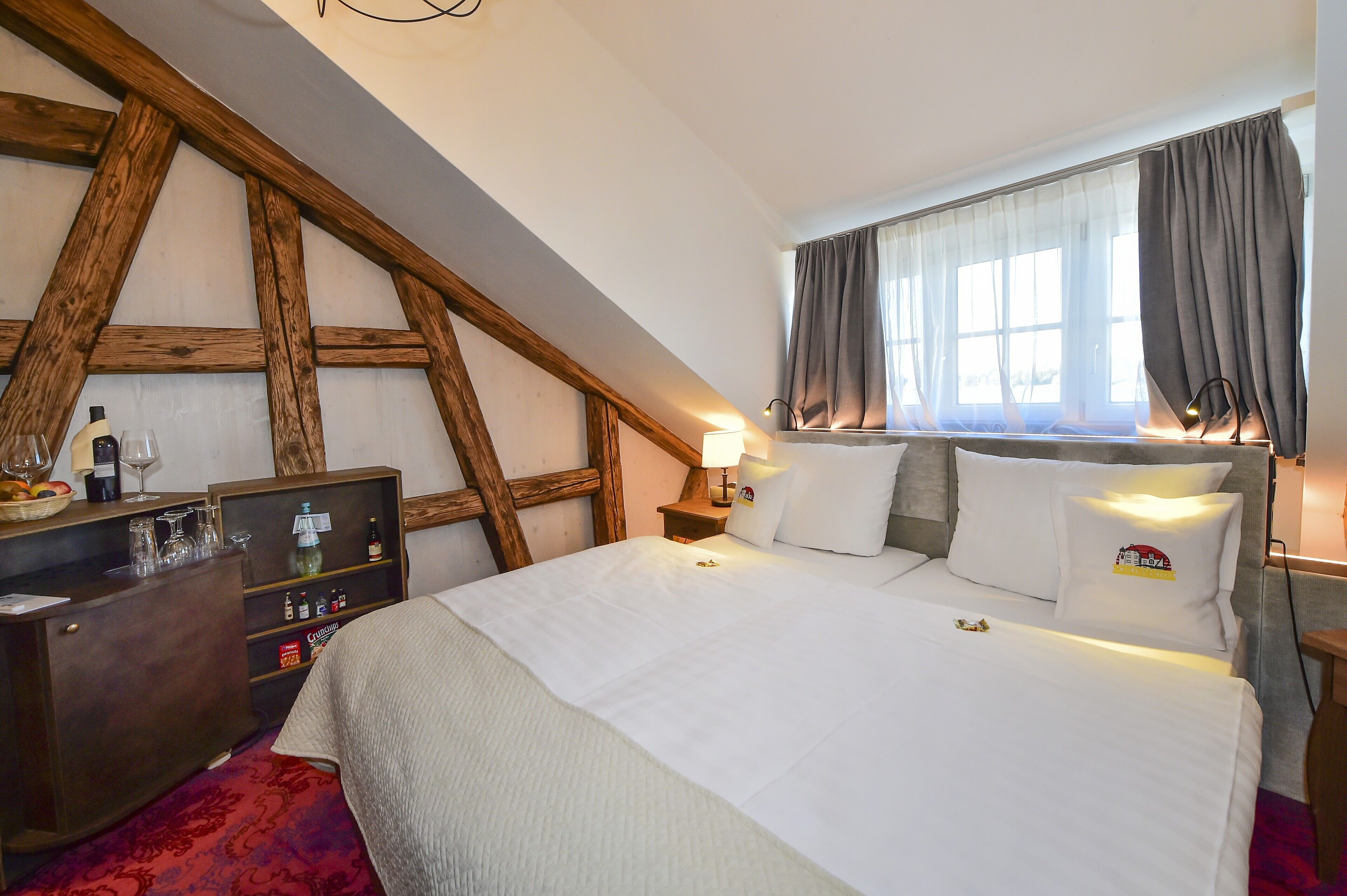Une chambre avec un plafond en pente, une commode en bois, un lit avec du linge de maison blanc et une fenêtre avec des rideaux.