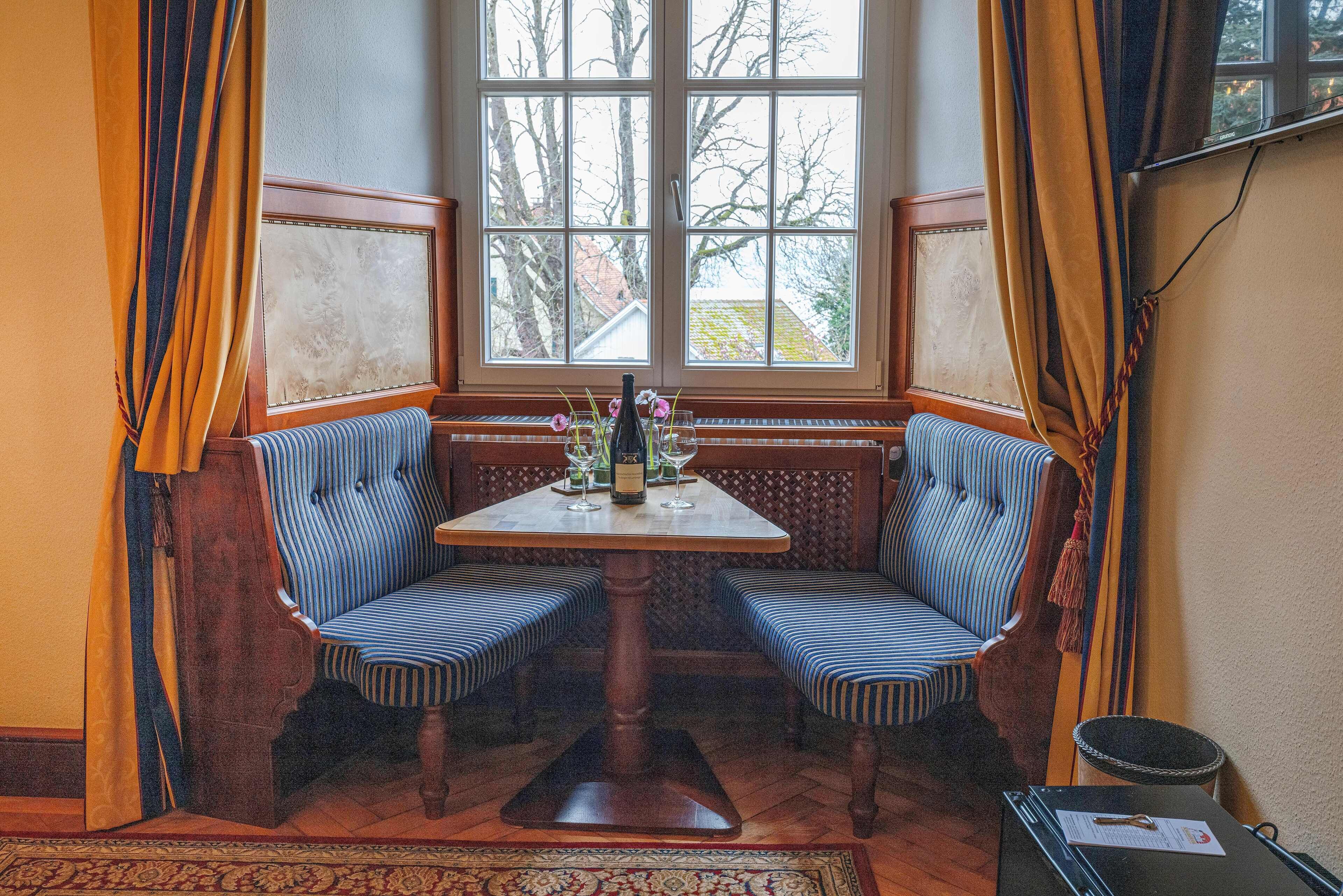 Eine Sitzecke mit Tisch und zwei gestreiften Stühlen, ein Fenster mit Vorhängen sowie ein kleiner Tisch mit Vase und Gläsern.