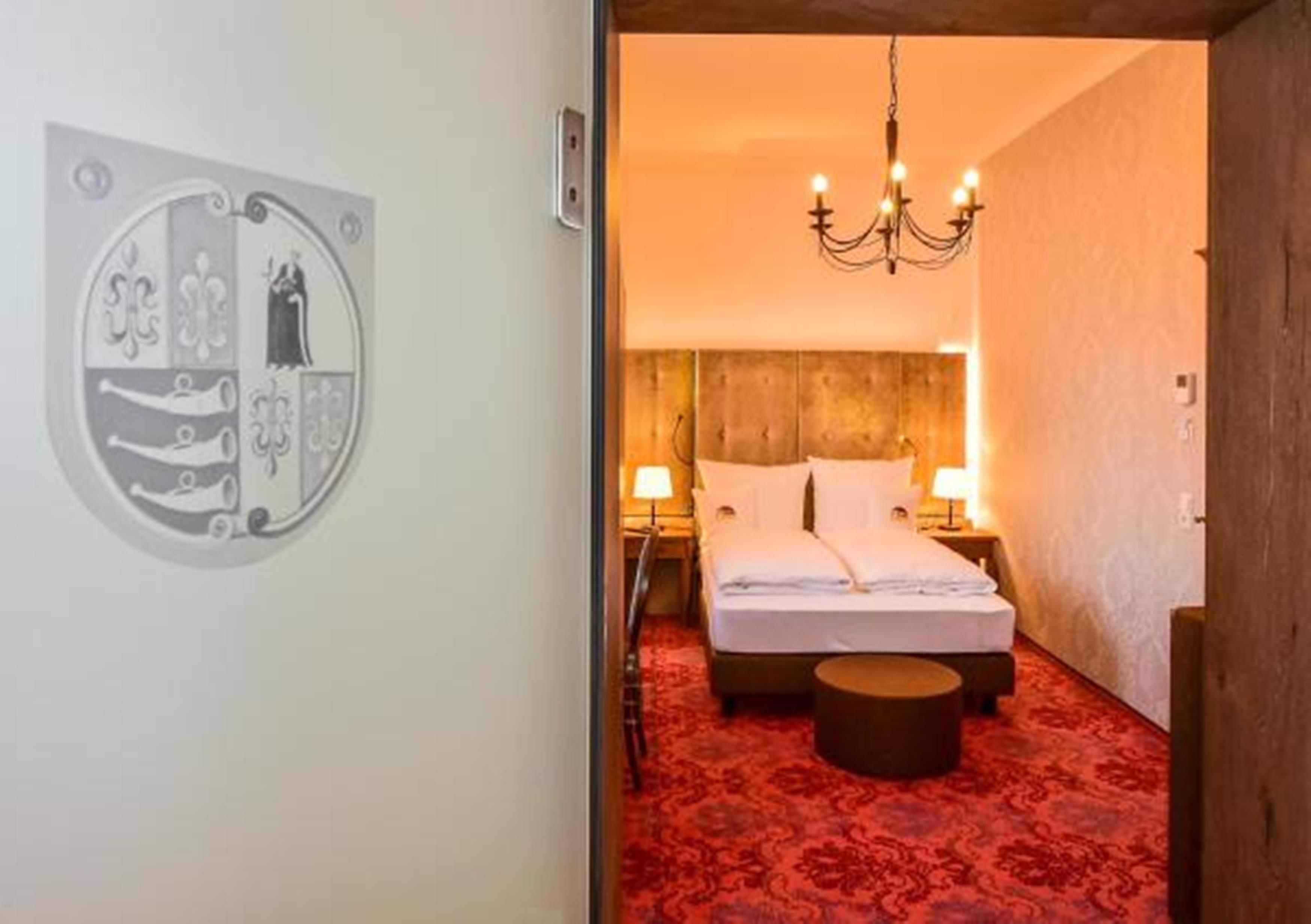 A hotel room with a bed, a round ottoman, a chandelier, and a wall-mounted artwork.
