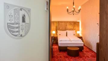 A hotel room with a bed, a round ottoman, a chandelier, and a wall-mounted artwork.