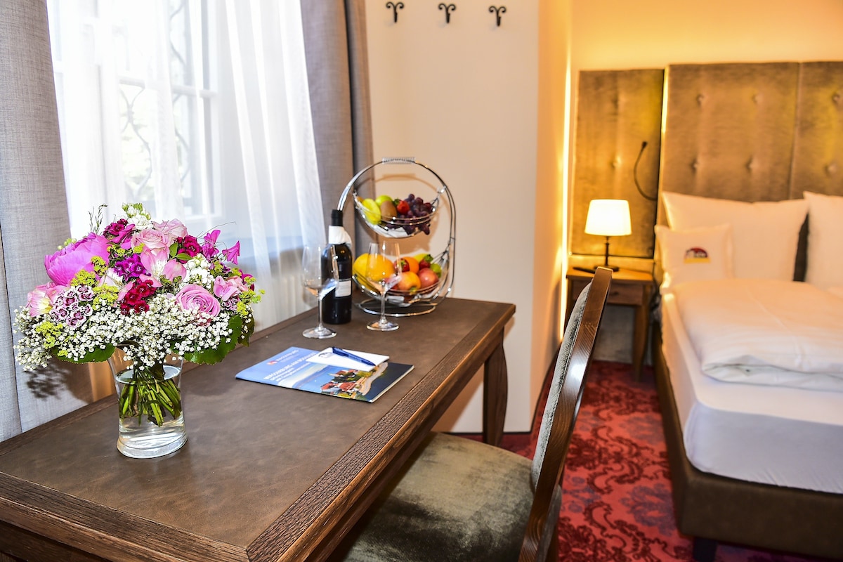 A hotel room with a wooden desk, a vase of flowers, a fruit bowl, and a bed with white linens.