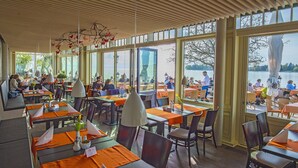 Un restaurante con mesas dispuestas para comer, amplias ventanas que dan a un lago y un techo de madera.