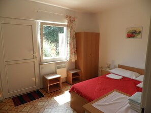 Ein Schlafzimmer mit Bett, Kleiderschrank, kleinem Tisch, einem Fenster mit Vorhängen und einer Heizung.