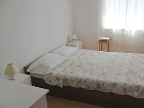 Une chambre simple et épurée, avec un lit, une table de chevet dotée d’une lampe et une petite commode.