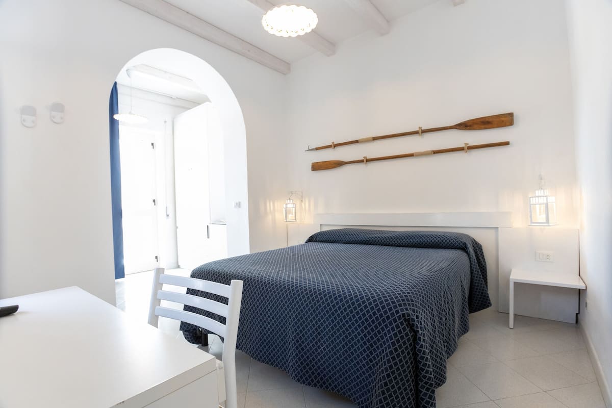 Quarto com cama, escrivaninha e cadeira. Há duas luminárias fixadas na parede e dois remos de madeira.