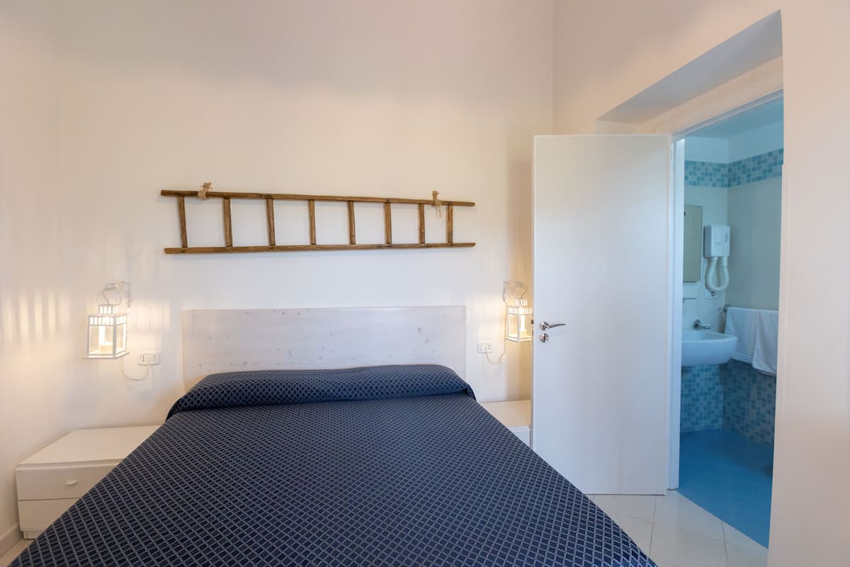 Quarto com cama, luminárias de cabeceira, prateleira de madeira e banheiro visível através de uma porta aberta.
