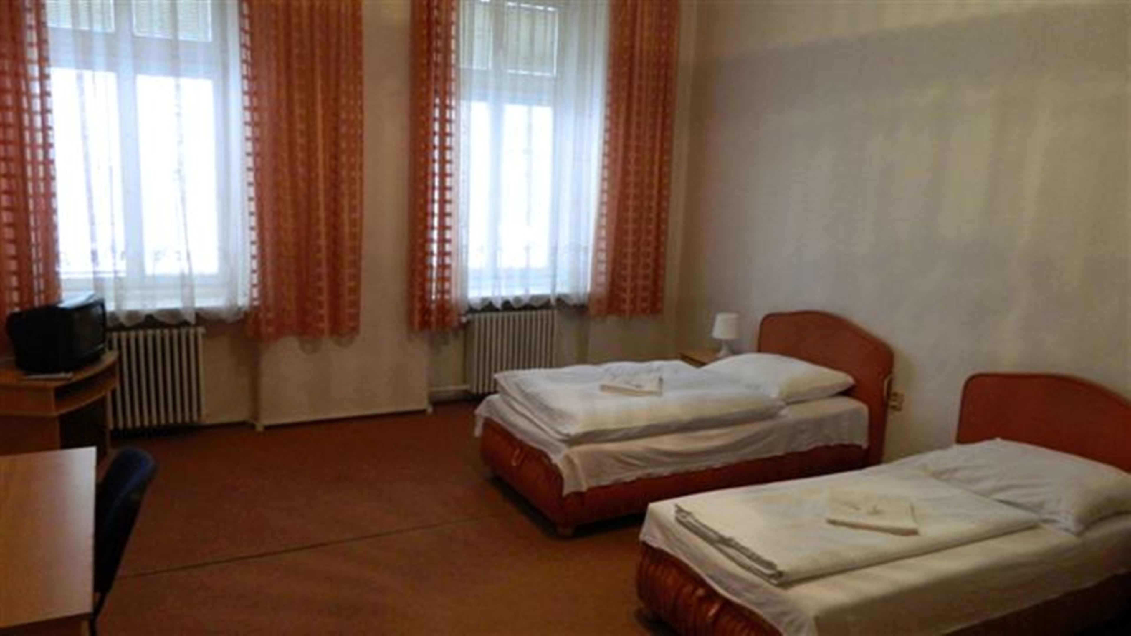 A hotel room with two single beds, a desk with a chair, a television, and a radiator.