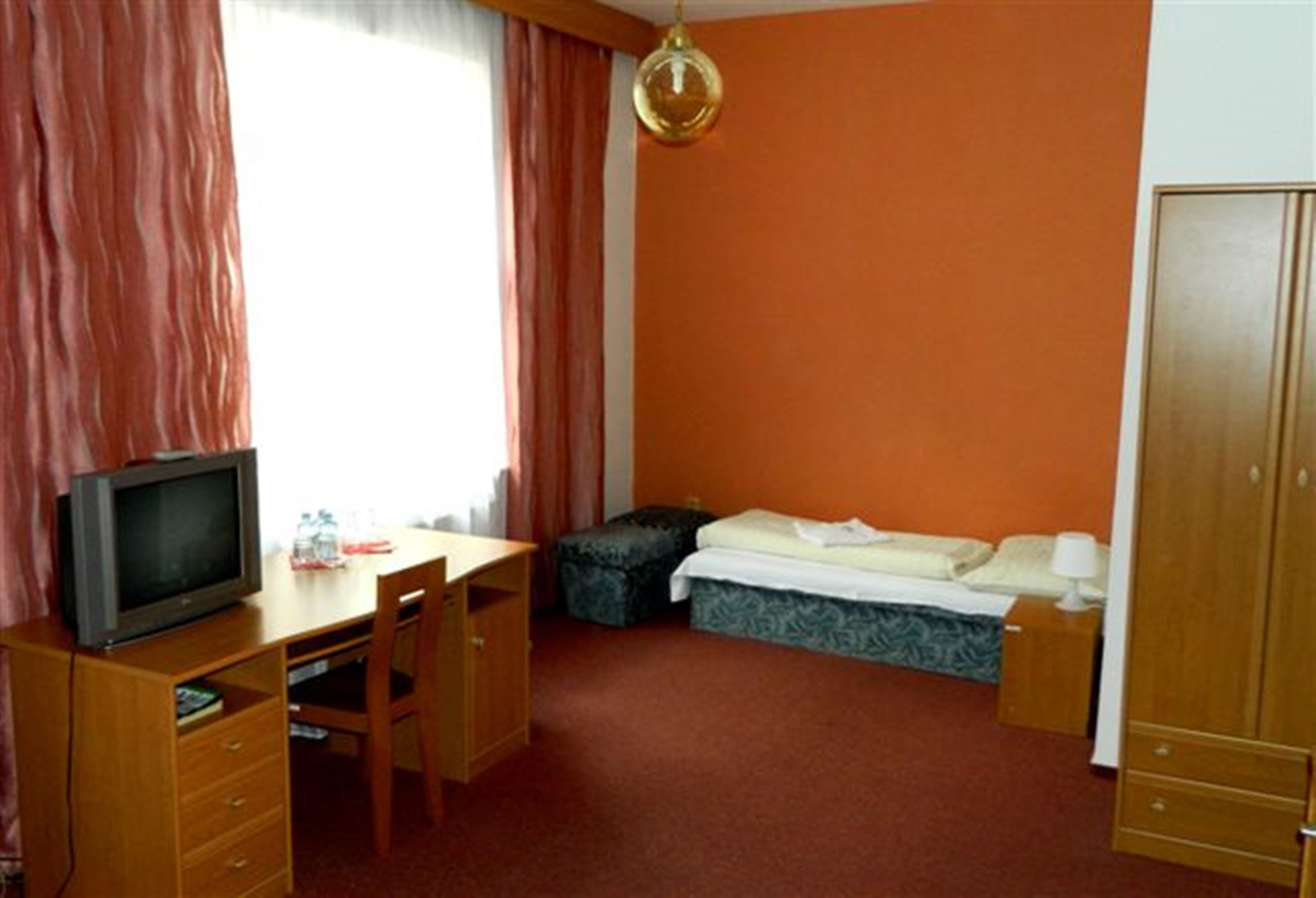 A small, single-room hotel room with a wooden desk, a television, a bed, and a wardrobe.