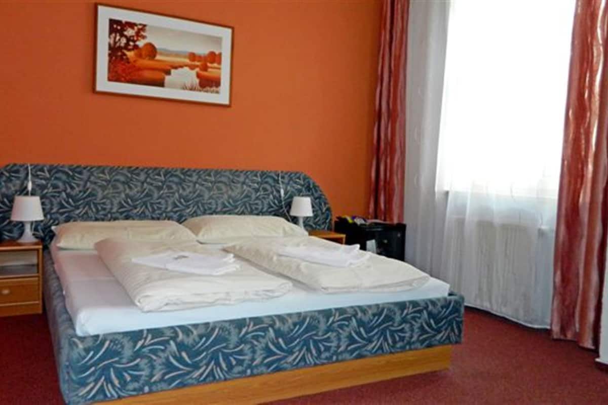 A hotel room with a bed, two bedside tables, a nightstand, a lamp, a painting on the wall, and a window with curtains.