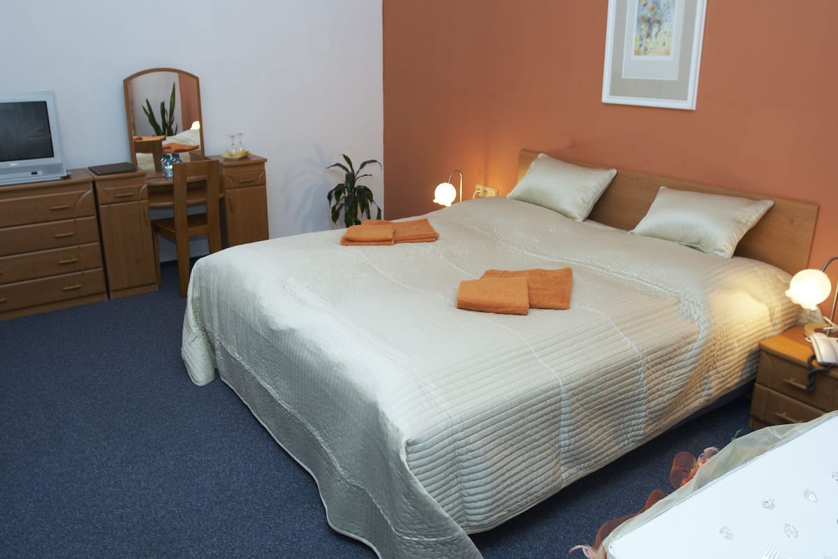 A neatly made bed with a white comforter and two orange towels, a wooden nightstand with a lamp, a desk with a computer and a mirror, and a potted plant.