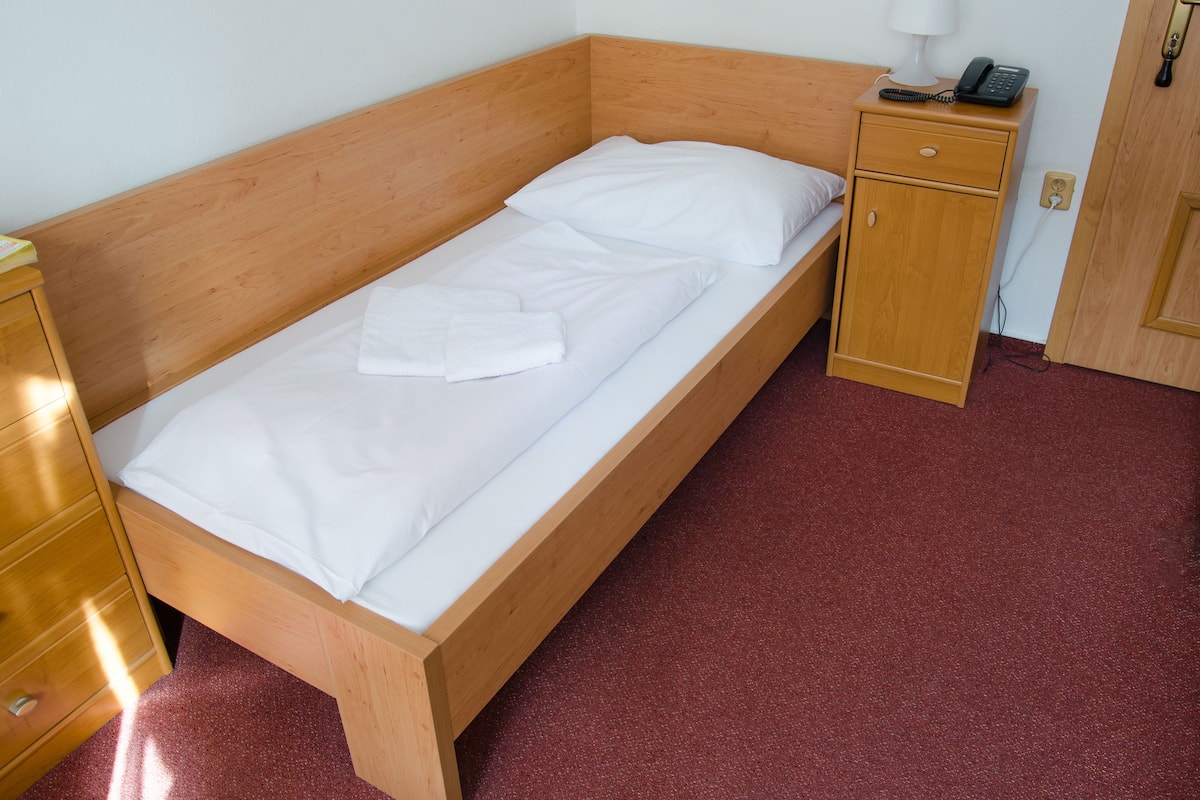 A single bed with a wooden frame, white bedding, and a bedside table with a lamp and phone.