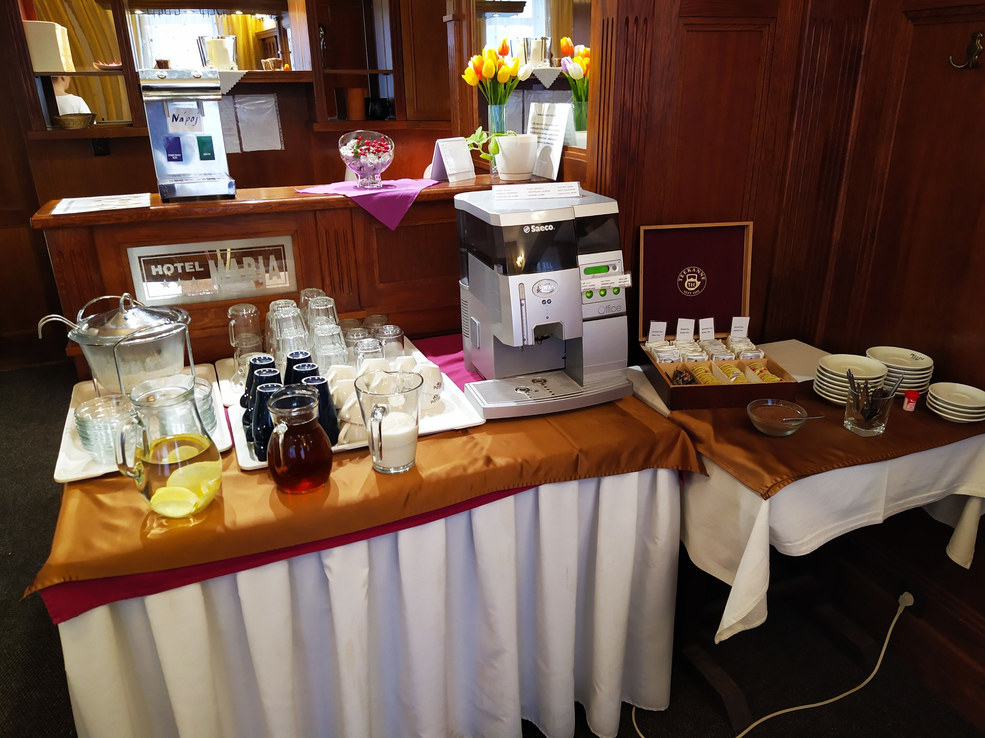 Uma área de recepção de hotel com máquina de café, espaço para chá e mesa de buffet com diversos alimentos e acompanhamentos.