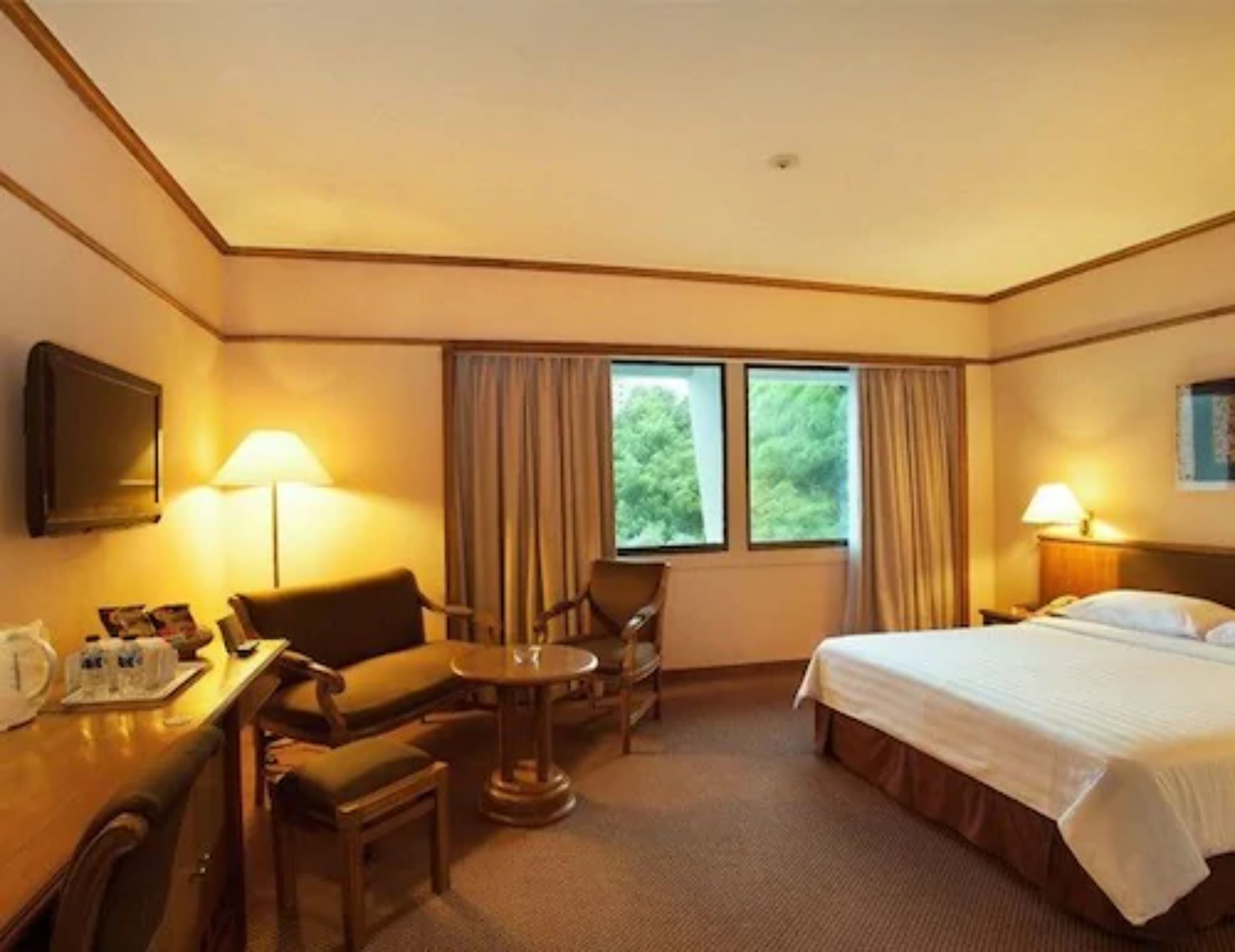 A hotel room with a bed, a desk with a chair, a small table, and two windows with curtains.